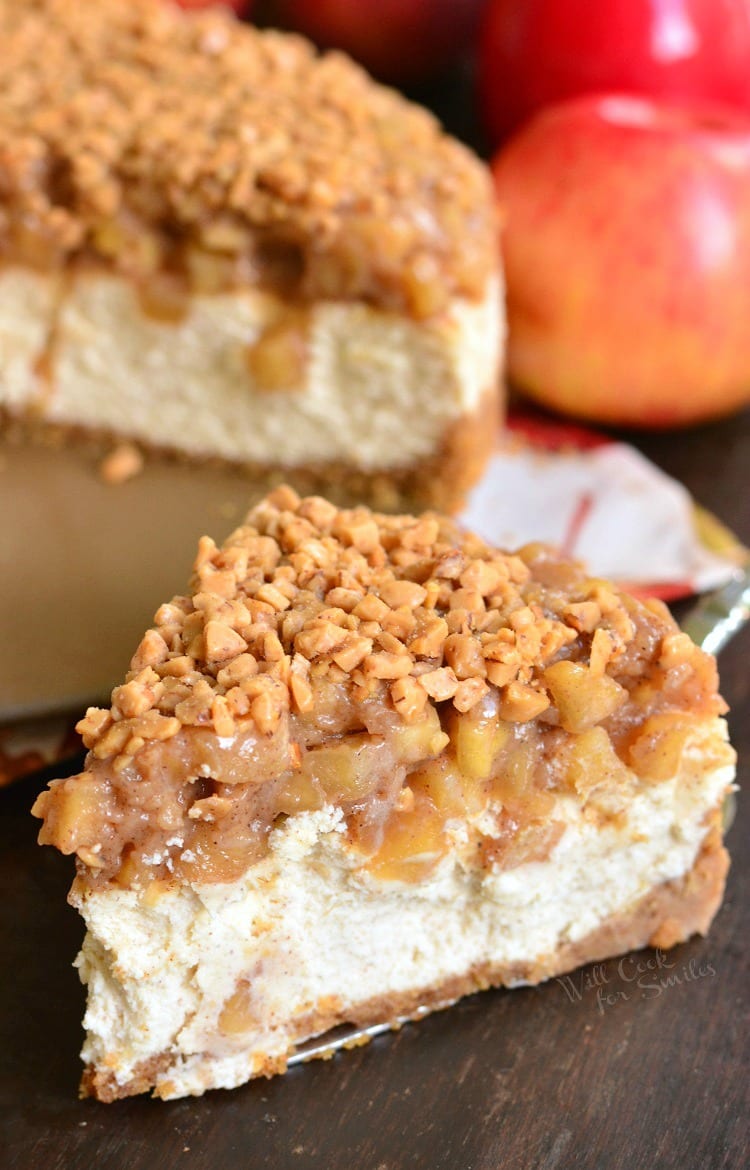 Apple Pie Cheesecake piece on a cutting board with the rest of the cheesecake in the background 