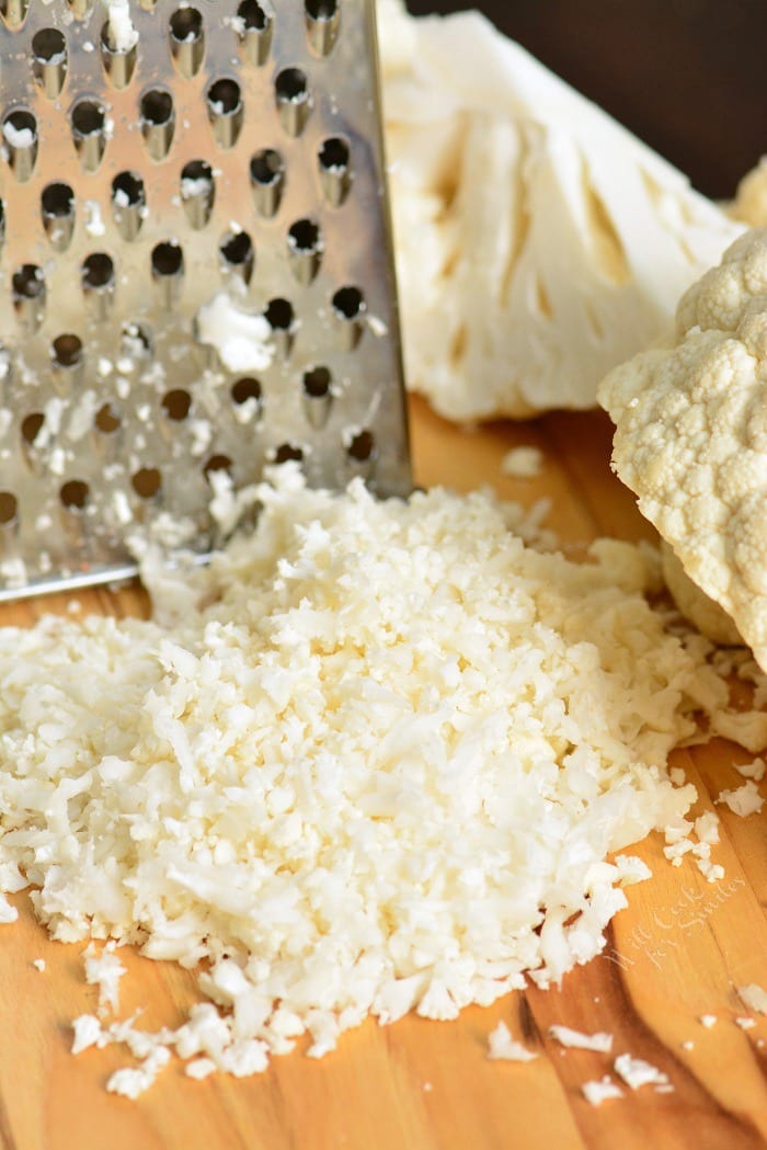 Cauliflower being shredded with grater onto a cutting board  