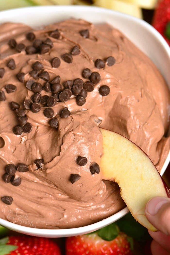 chocolate cake dip in a white bowl with chocolate chips on top and a apple slice being dipped in the bowl.