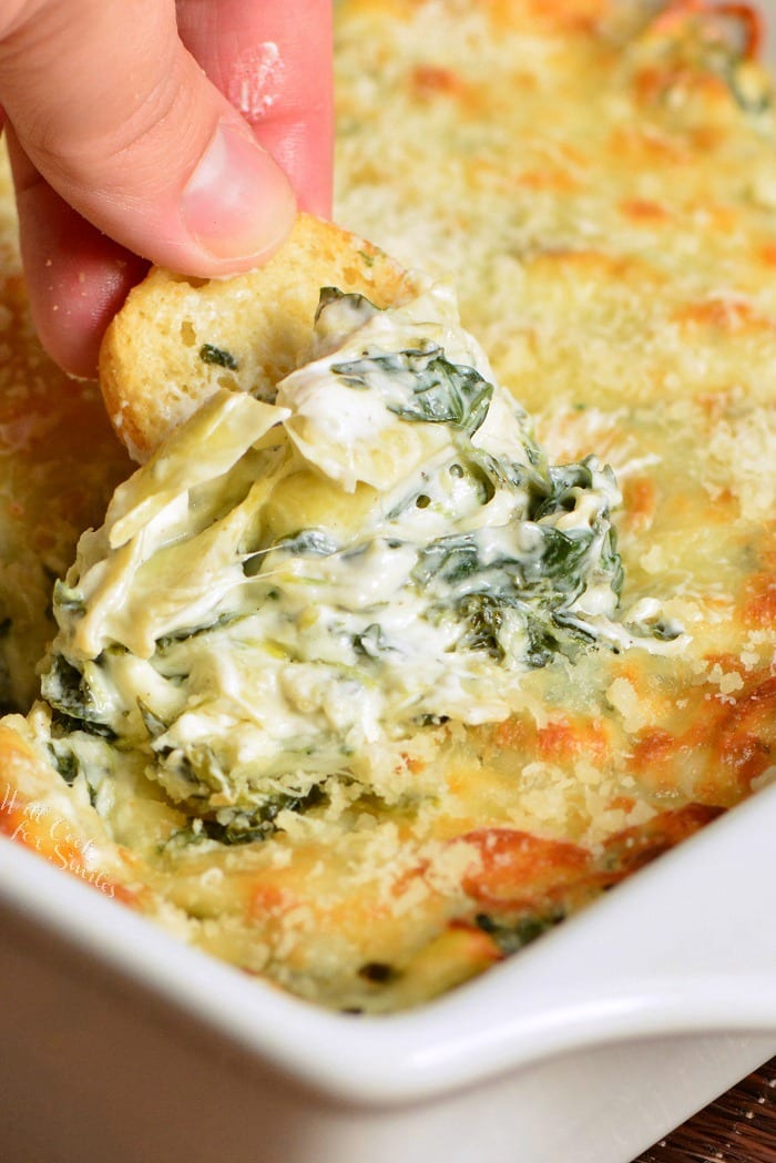 dipping bagel chip in Spinach Artichoke Dip in a white baking dish 