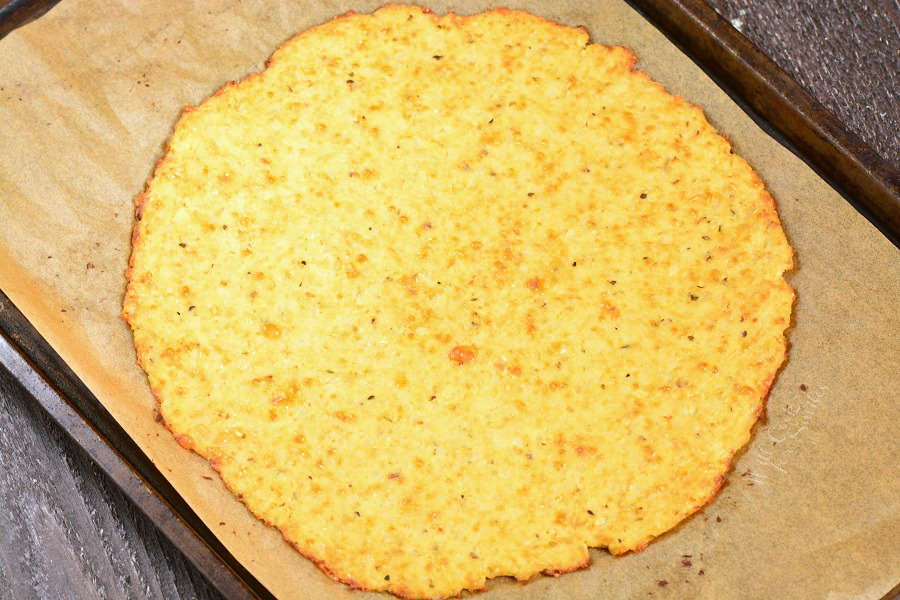 Cauliflower Pizza Crust cooked on parchment paper on a baking sheet sitting on a wood cutting board 