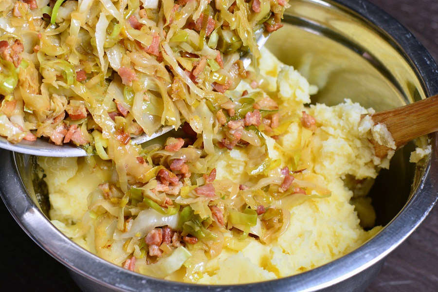 cabage and bacon mixture being poured out of pan into metal mixing bowl with mash potatoes and stirred with wooden spoon 