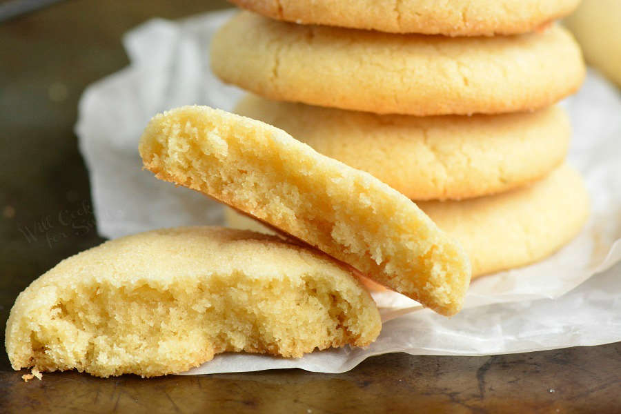 Sugar Cookies are stacked up and one cut in half on wax paper on a wood cutting board 