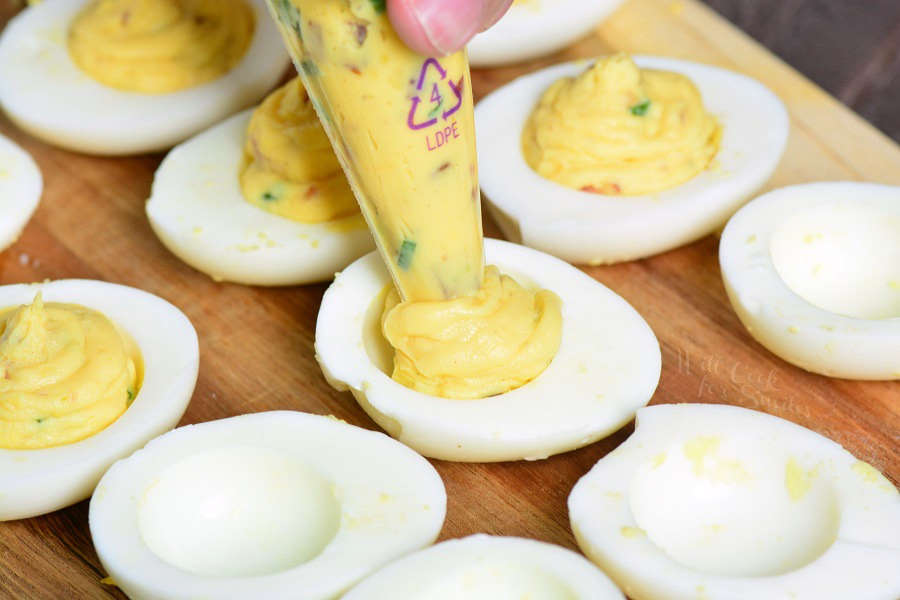 deviled egg filling in a piping bag being piped into the egg halves that are sitting on a cutting board 