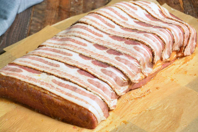 preparing brisket by wrapping bacon around brisket, sitting on parchment paper on a wood cutting board 