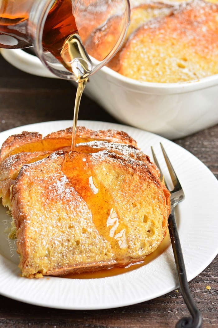 baked french toast on a plate with a fork with syrup being poured over it on a wood table 
