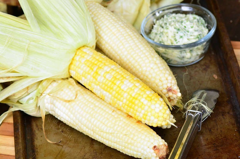 grill corn with husks pealed back, with a bowl of herb butter, and tongs on a baking sheet 
