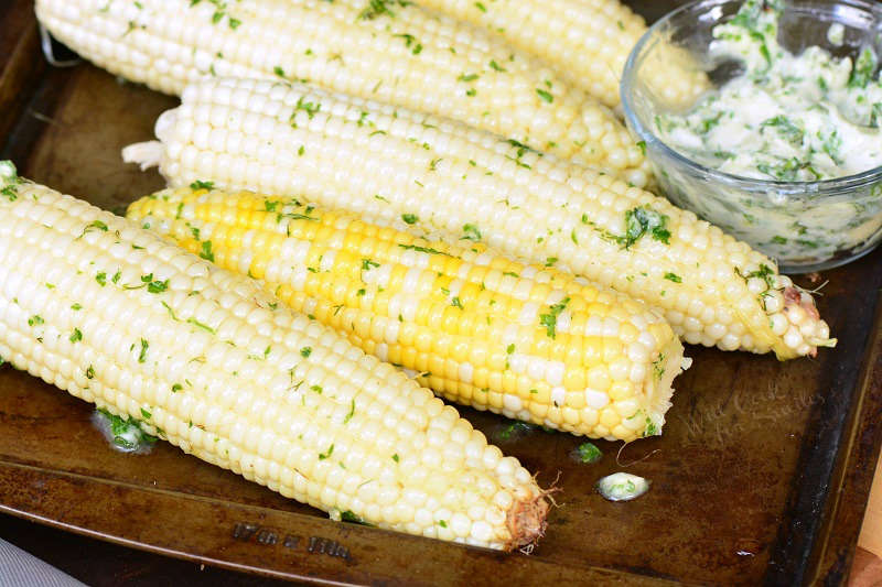 Corn on the cub on a baking sheet with a bowl of herb butter 