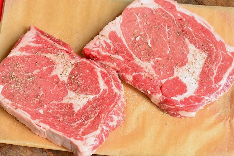 raw rib eye steaks on parchment paper on top of a wood cutting board 