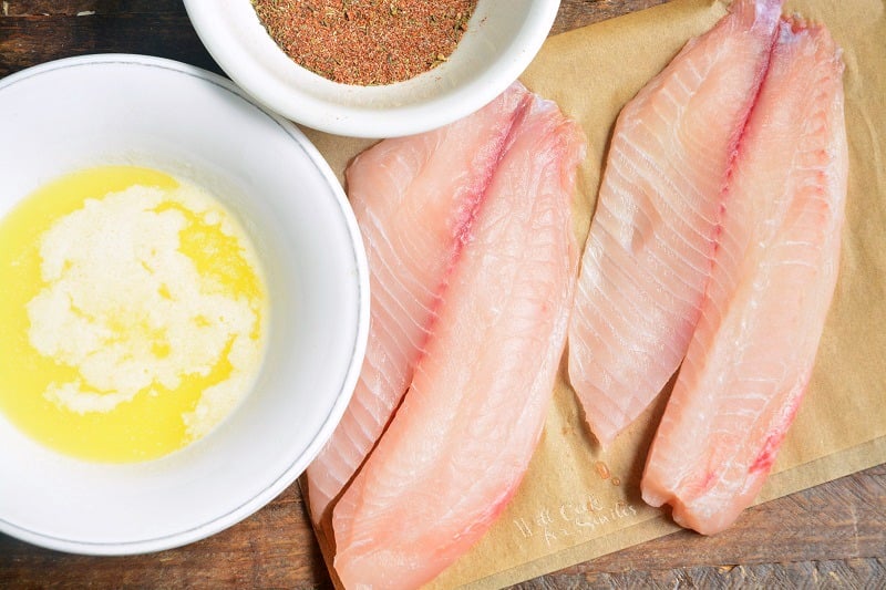 melted butter in a bowl, seasoning in a bowl, and fish on parchment paper on a wood cutting board 