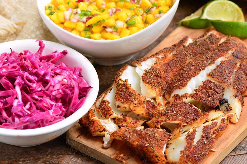 fish on a wooden cutting board, red cabbage in a bowl and mango salsa in a bowl 