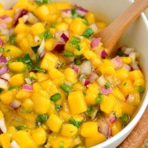 Mixed mango salsa in a white bowl with a wooden spoon.