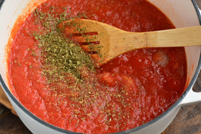 crushed tomatoes in a white Dutch oven with seasonings and s wooden spoon on top