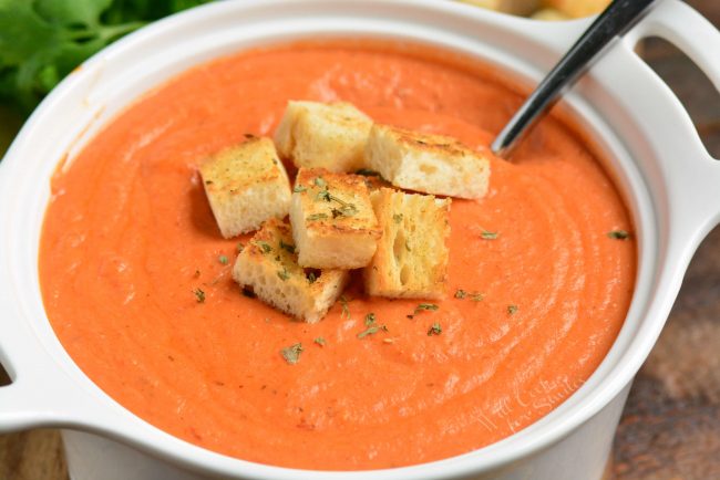 closer horizontal view of the tomato bisque in a white bowl with croutons on top and a spoon in a bowl
