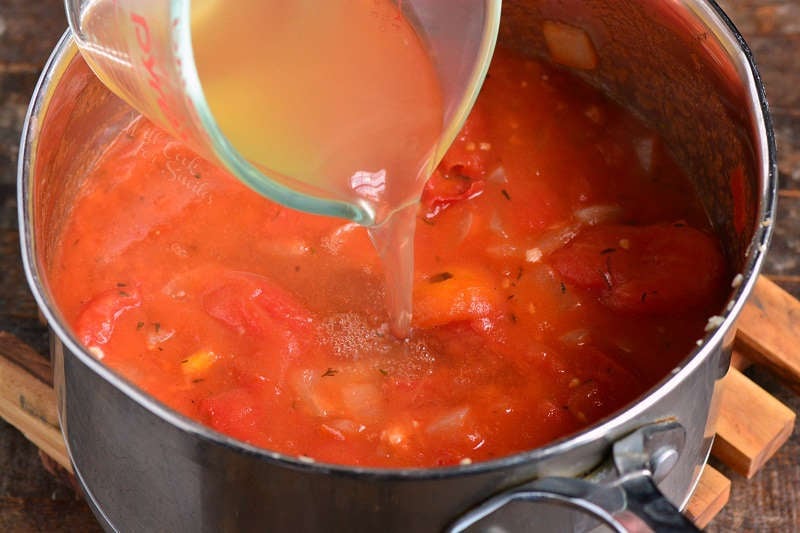 pouring chicken stock into the soup pot with tomatoes and other ingredients cooking