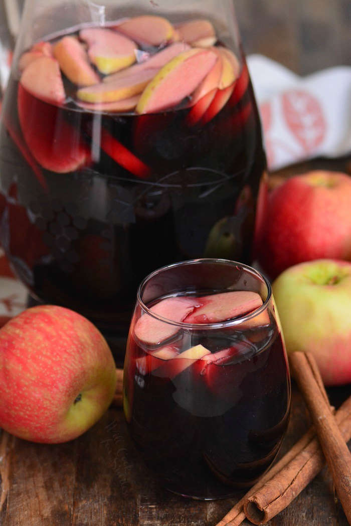 Fall apple sangria in a pitcher and poured into a wine glass on a wood table with apples and cinnamon 