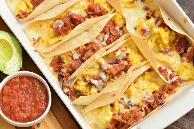 baked breakfast tacos in a baking pan with a bowl of salsa and some avocado on a wood cutting board 