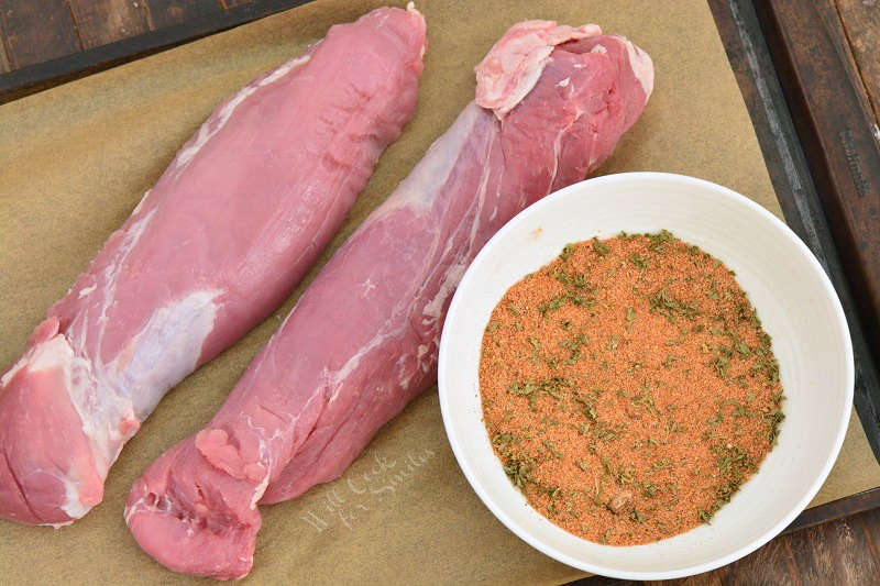 two pork tenderloin and seasoning on a piece of butcher paper on a backing sheet that is on a wood table 