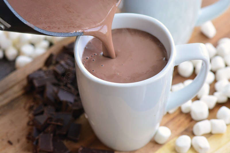 pouring hot chocolate into a mug on a cutting board with chocolate and marshmallows 