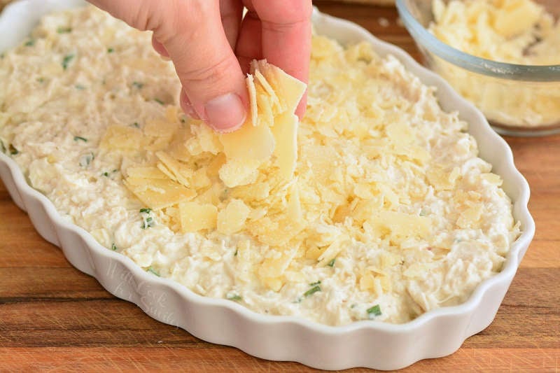 putting cheese on top of a crab dip in a glass serving bowl on top of a wood cutting board 