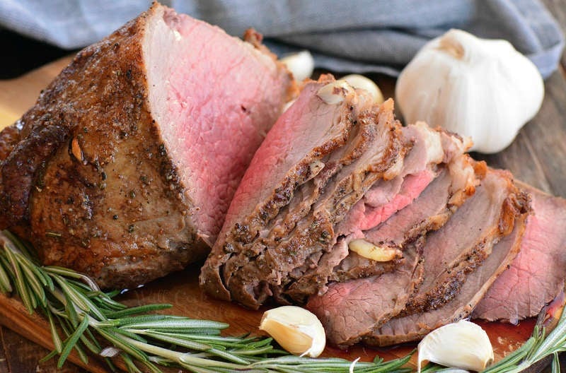 sliced roast beef horizontal on a wood cutting board with a sprig of rosemary and garlic cloves 