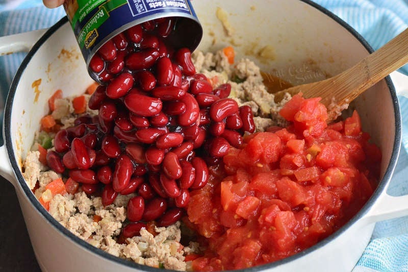 pouring in red kidney beans out of a can into a pot with cooked turkey vegetables and diced tomatoes