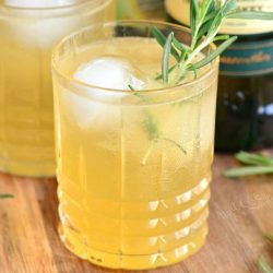 closeup of light yellow cocktail with ice and rosemary.