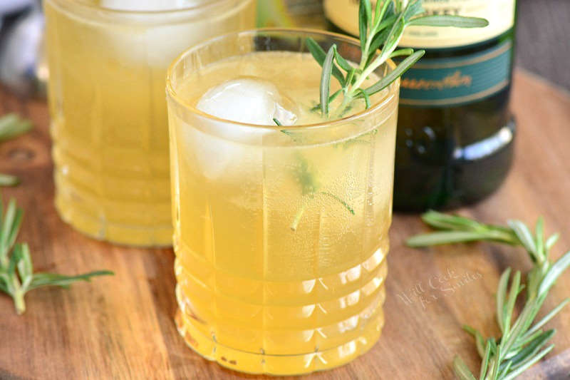 closeup of light yellow cocktail with ice and rosemary.
