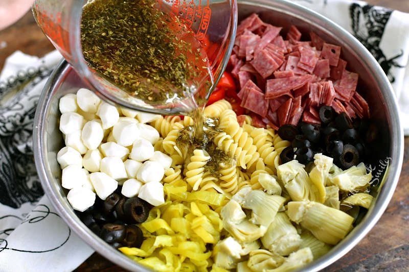 pouring dressing over Antipasto Pasta Salad in a metal bowl 