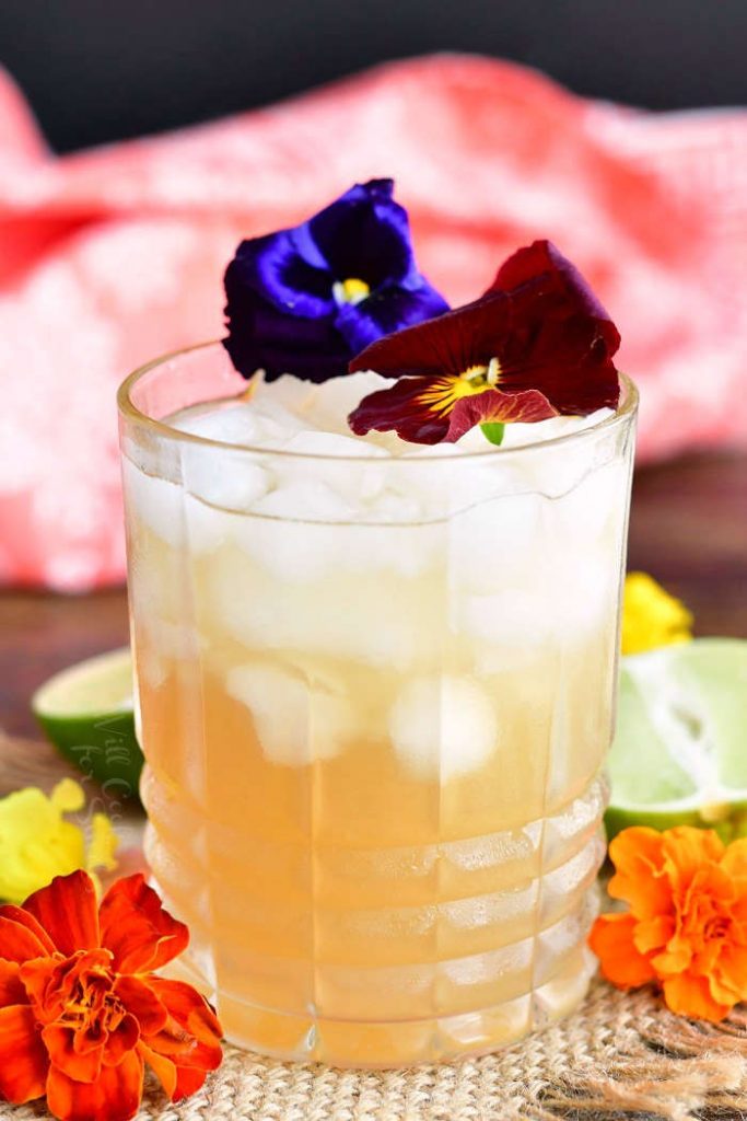 closeup of glass filled with crushed ice and light yellow cocktail decorated with purple and burgundy flowers