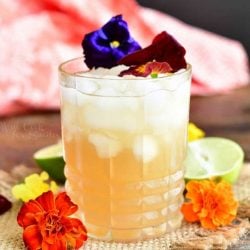 horizontal side view of the yellowish cocktail in a glass topped with two flowers and more orange flowers around the glass
