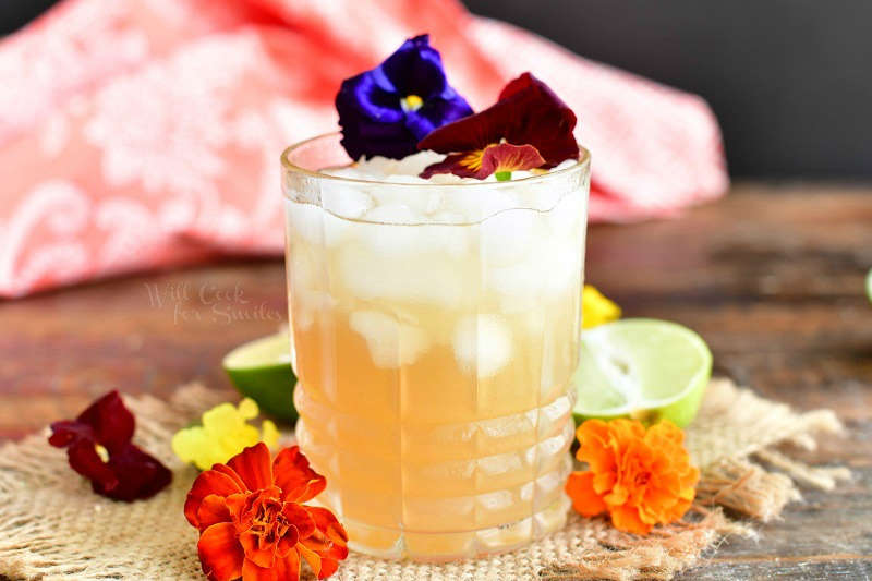 horizontal side view of the yellowish cocktail in a glass topped with two flowers and more orange flowers around the glass