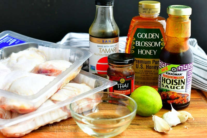 ingredients for baked chicken thighs on a cutting board including 2 packages of chicken thighs, soy sauce, honey, hoisin sauce, chili paste, lime, garlic cloves, and vinegar.