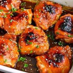 horizontal closer view of baked chicken thighs in dark brown sauce in a white baking dish with green onions on top and sesame seeds