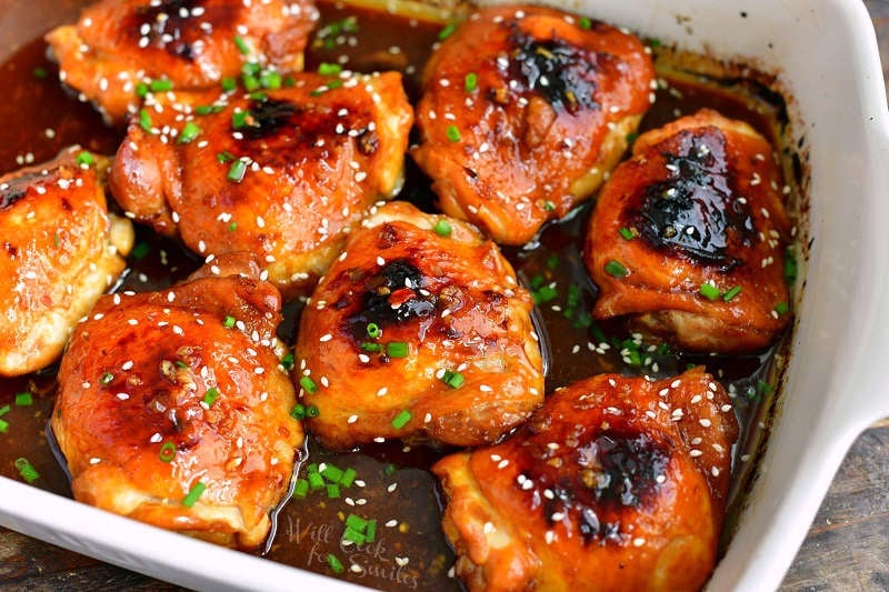 horizontal closer view of baked chicken thighs in dark brown sauce in a white baking dish with green onions on top and sesame seeds