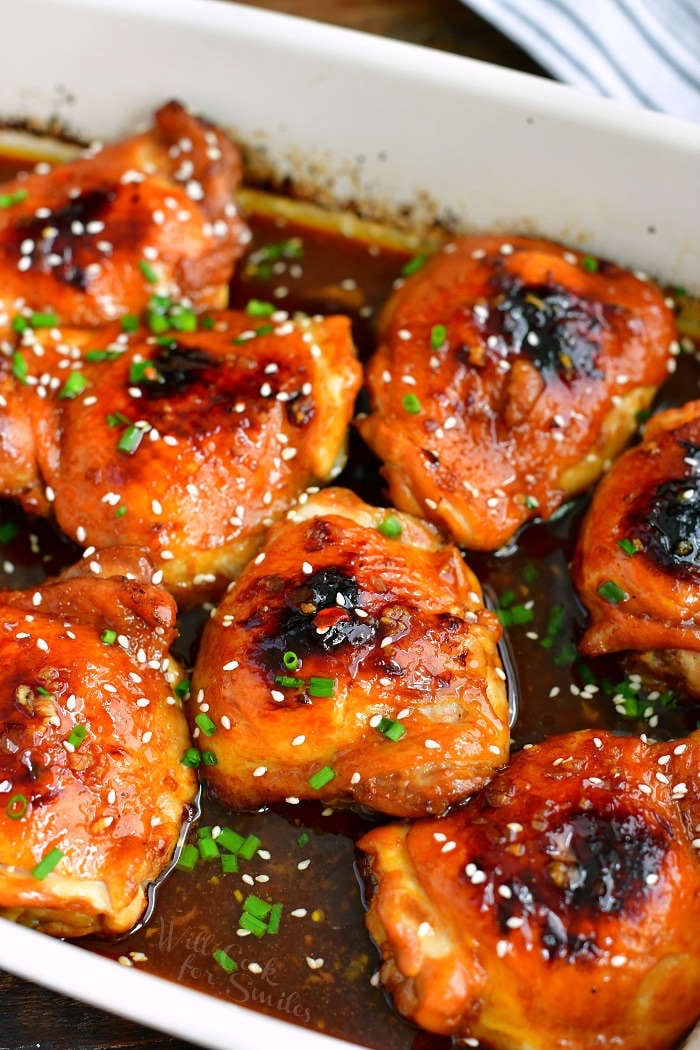 baked chicken thighs in dark brown sauce in a white baking dish with green onions on top and sesame seeds