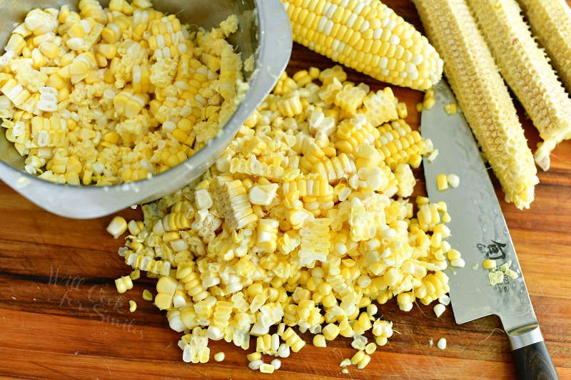 corn on a cob and corn kernels cut off the cob on the cutting board with a knife and a bowl of some corn kernels in it