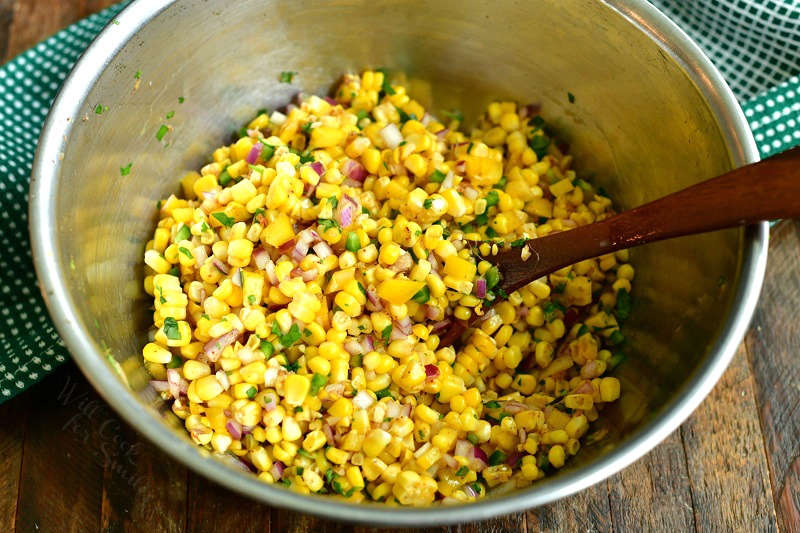 ingredients for corn salsa mixed together in a metal mixing bowl with a wooden spoon