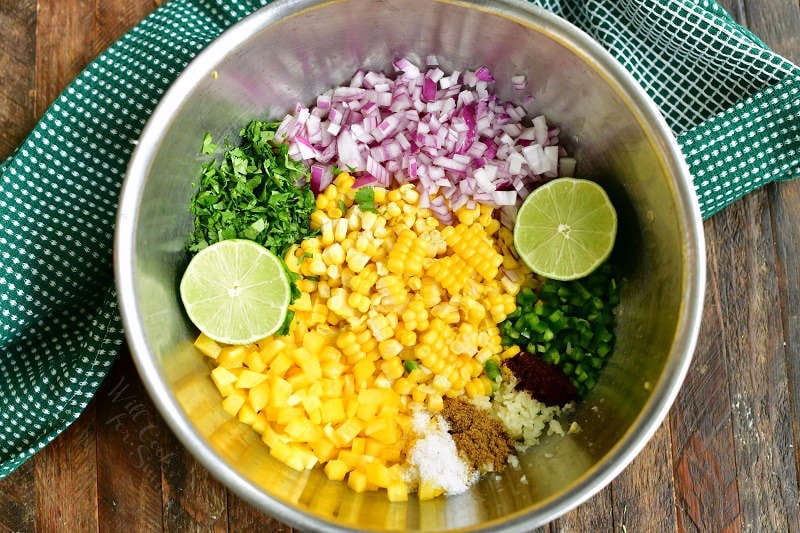 ingredients in a bowl side by side including corn kernels, yellow bell pepper, lime halves, jalapeno, red onion, cilantro, and seasonings