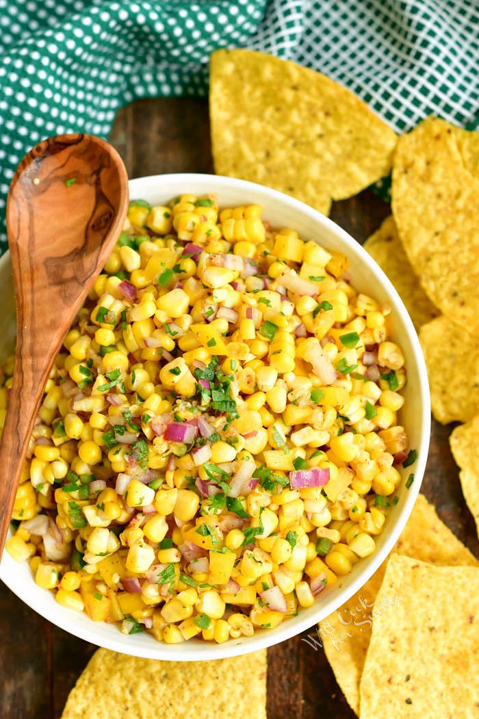 top view of corn salsa in a white bowl with wooden spoon laying on the bowl and chips around it