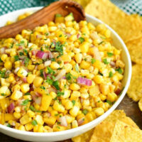 horizontal image of corn salsa in a white bowl with wooden spoon in it and yellow corn tortillas around