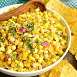 horizontal image of corn salsa in a white bowl with wooden spoon in it and yellow corn tortillas around