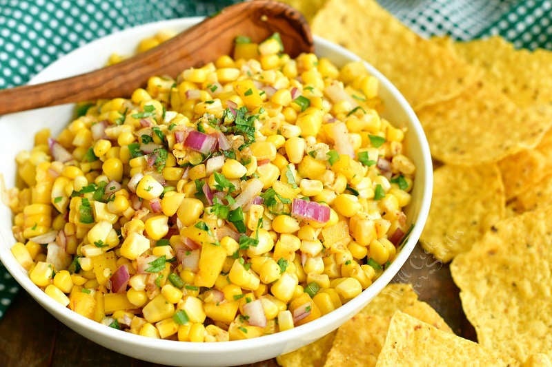 horizontal image of corn salsa in a white bowl with wooden spoon in it and yellow corn tortillas around