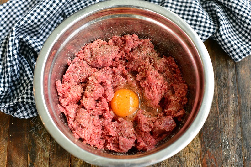 ground beef in a mixing bowl with an egg in the center and salt and pepper sprinkled on top