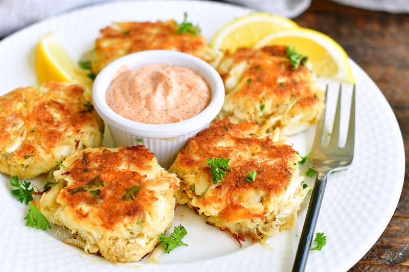 five crab cakes on a white place in a circle with cup of remoulade in the center and lemon wedges