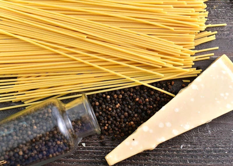 dried thin long pasta, whole peppercorns spilling out of a clear glass jar, and a block of hard cheese on a wooden board