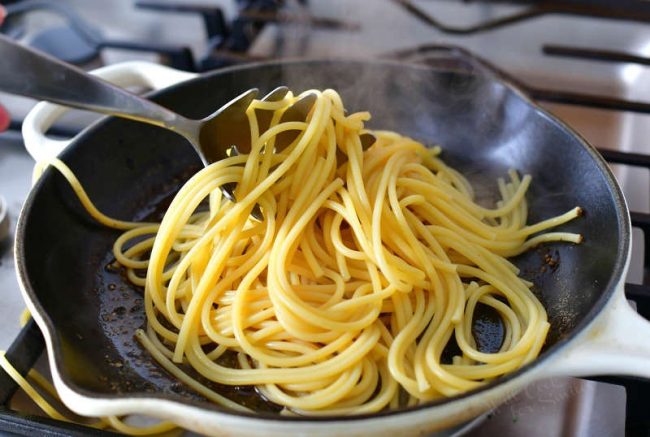 adding cooked pasta into the skillet on the stove with a pasta fork