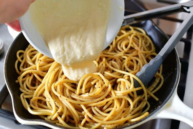 adding cheese sauce out of a white bowl into the pan with pasta mixed with pepper