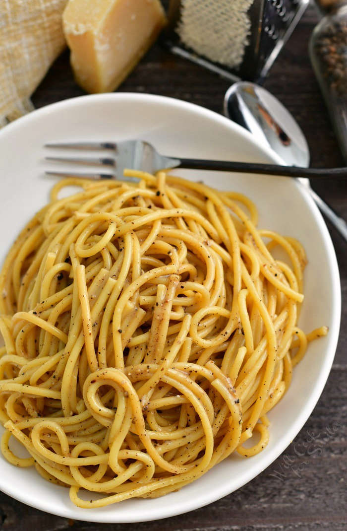 top view of cheesy and peppery pasta swirled in a white bowl with a fork in the bowl and spoon and block of cheese next to the bowl