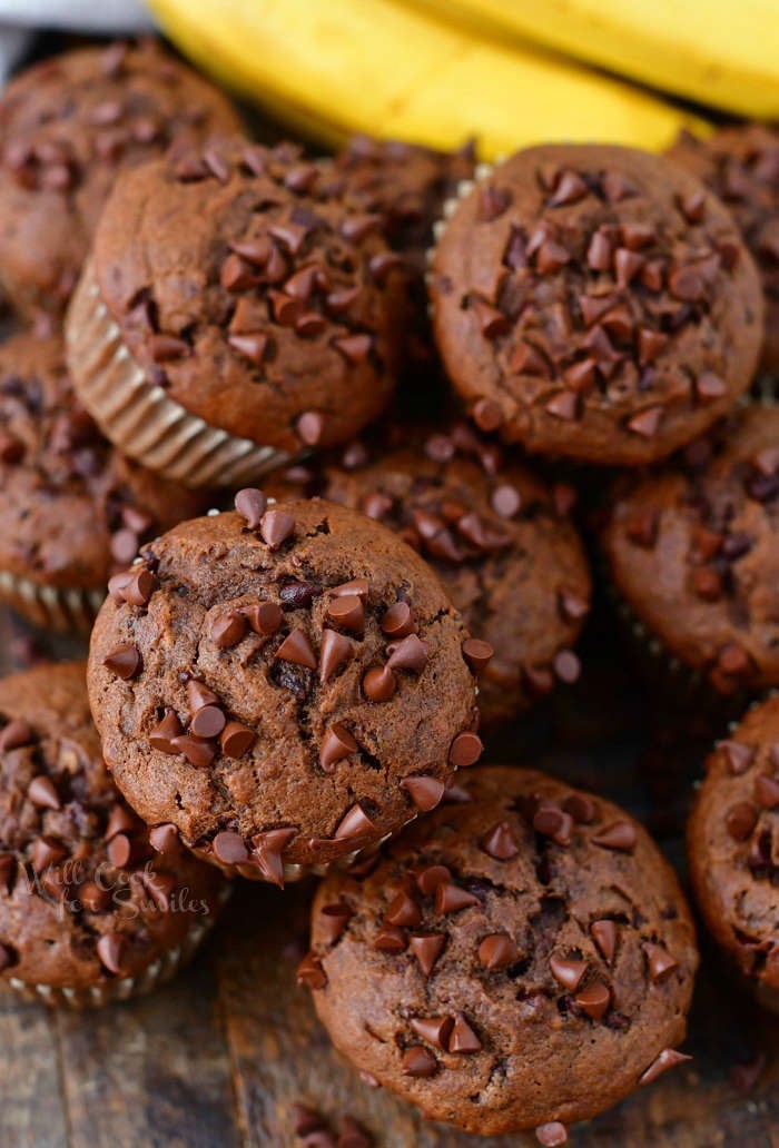 close up of chocolate muffins topped with mini chocolate chips on top of one another and some bananas on the background